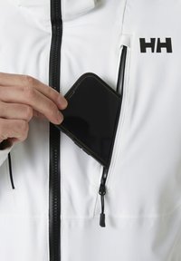 Bílá bunda s černým zipem a kapsou na zip na prsou, do které ruka vkládá černý smartphone. Na bundě je viditelné logo.