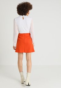 Blusa blanca de manga larga con detalle de abertura en la parte posterior, combinada con una falda acampanada de color naranja brillante. Calzado: botines crema con tacón.