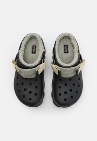 Zuecos negros con forro gris suave, orificios de ventilación y correa ajustable beige, presentando una base de goma sólida y un detalle de logo.