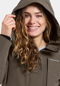 Uśmiechnięta kobieta z kręconymi włosami, ubrana w brązową kurtkę z kapturem Didriksons, trzymająca kaptur jedną ręką.