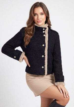 Kvinde med langt bølget hår iført en sort tekstureret jakke med beige pelskant og knapper, beige nederdel, poserer med den ene hånd på hoften.