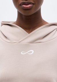 Gros plan d'une personne portant un sweat à capuche beige avec un symbole infini blanc sur la poitrine, montrant uniquement les lèvres et le cou.