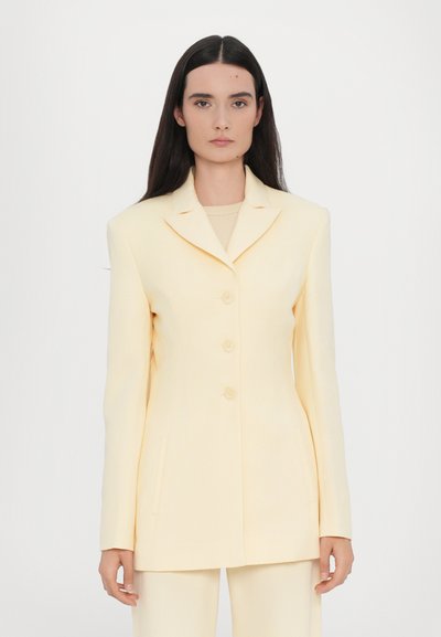Yellow tailored blazer with a notch lapel, four buttons, and side pockets. Smooth fabric, structured silhouette, and long sleeves.