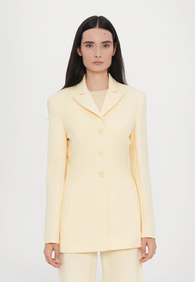 Yellow tailored blazer with a notch lapel, four buttons, and side pockets. Smooth fabric, structured silhouette, and long sleeves.
