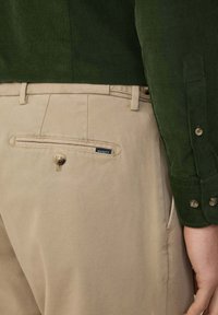 Beżowe spodnie o gładkiej fakturze, z tylną kieszenią z zapięciem na guzik, zestawione z ciemnozieloną koszulą z miękkiego materiału.