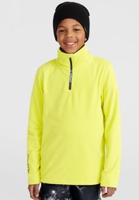 Pull en polaire jaune vif avec un col haut, une fermeture éclair et des manches longues. Logo sur la manche gauche. Porté avec un pantalon à motifs sombres.
