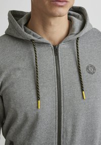 Grijze zip-up hoodie met een gestreept patroon, voorzien van een capuchon met trekkoord en gele uiteinden, en een geborduurd logo op de borst.