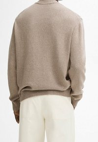 Homme portant un pull en tricot taupe et un pantalon écru, vu de dos, se tenant devant un fond blanc uni.