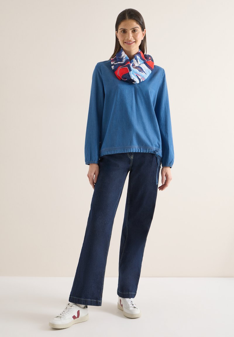 Femme debout souriante, portant un haut bleu à manches longues, un jean foncé, des baskets blanches et une écharpe colorée rouge, blanche et bleue.