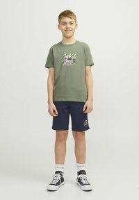 Camiseta de algodón verde oliva con un gráfico en blanco y púrpura de "Jack & Jones", combinada con pantalones cortos azul marino con un logo. Zapatillas negras y calcetines blancos.
