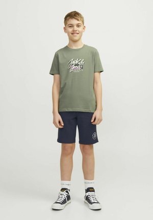 Camiseta de algodón verde oliva con un gráfico en blanco y púrpura de "Jack & Jones", combinada con pantalones cortos azul marino con un logo. Zapatillas negras y calcetines blancos.