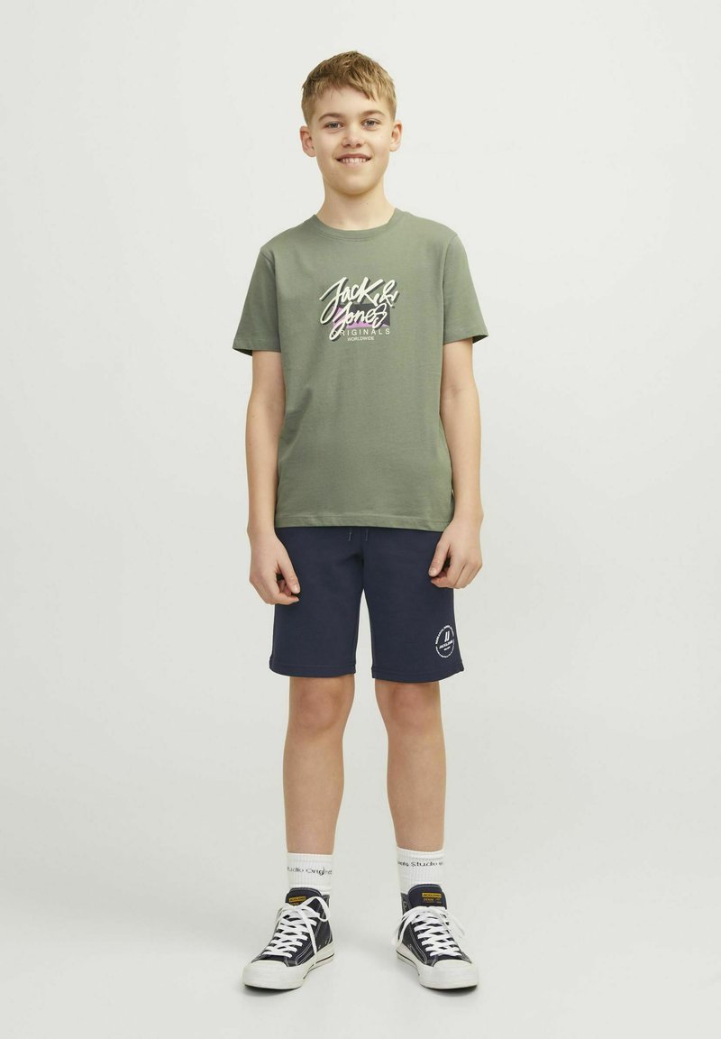 Camiseta de algodón verde oliva con un gráfico en blanco y púrpura de "Jack & Jones", combinada con pantalones cortos azul marino con un logo. Zapatillas negras y calcetines blancos.
