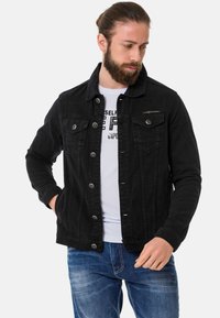 Veste en denim noir avec un devant à boutons, deux poches poitrine et un ourlet légèrement effiloché. Portée sur un t-shirt graphique blanc.