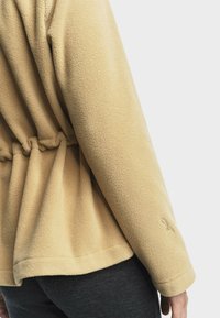 Veste en polaire beige avec un design de dos froncé, tissu texturé et un subtil détail brodé sur la manche.