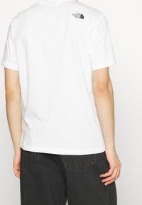 Person som bär en vit t-shirt med kort ärm och en liten svart "The North Face"-logotyp på övre ryggen, kombinerad med svarta jeans.