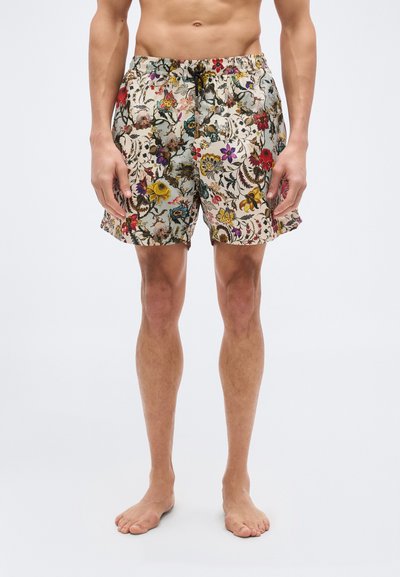 Torso et jambes d'un homme portant un short à cordon à motif floral, debout pieds nus sur un fond blanc uni.