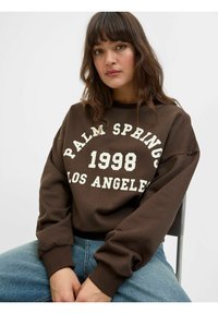 Felpa marrone con la scritta "PALM SPRINGS 1998 LOS ANGELES" in bianco. Presenta un collo rotondo e polsini a costine, abbinata a jeans in denim blu.
