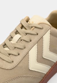 Beige suede sneaker with cream-colored leather accents, zigzag design, and textured laces. Rubber sole and contrasting detailing visible.
