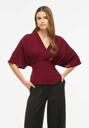 Femme aux longs cheveux portant un haut bordeaux ajusté à manches larges et un pantalon noir taille haute, debout les mains dans les poches.