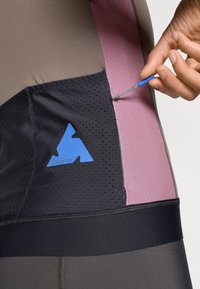 Abbigliamento sportivo caratterizzato da una combinazione di tessuti marroni, rosa e neri. Include un logo blu e un dettaglio con coulisse in una tasca.