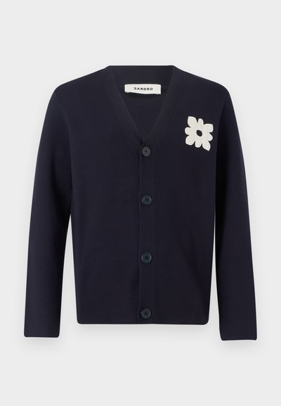 Tamsiai mėlynas mezginys kardiganas su V-formos iškirpte, ilgomis rankovėmis ir penkiais mygtukais. Kairiajame šone yra baltas gėlių aplikacija. Lygus paviršius.