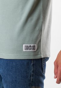 Ljusgrön strukturerad t-shirt med ett ribbat mönster, med en etikett vid fållen som säger "DENIM FACTORY," tillsammans med blå jeans.