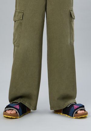 Pieds portant des sandales à bout ouvert avec des brides bleues et des semelles jaunes, associées à un pantalon cargo ample vert olive.