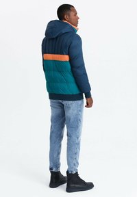 Puffjacka med en marinblå överdel, teal nedre del och ett orange horisontellt band. Tillverkad av quiltat material med huva.