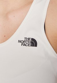 Bílé sportovní tílko vyrobené z elastického materiálu, s černým logem "THE NORTH FACE" na levé straně, s klasickým kulatým výstřihem.
