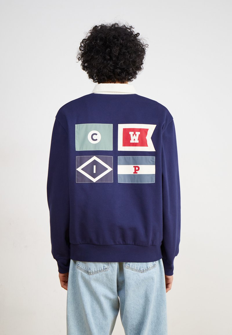 Marinblå sweatshirt med rund hals och flerfärgade tryckta lappar på baksidan, inklusive bokstäver och former i röd, grön och vit.