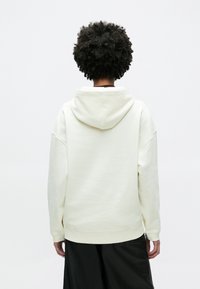 Világos bézs kapucnis pulóver nagy kapucnival, leengedett vállakkal és bordázott mandzsettákkal. Sima textúra, előtérben sötét szoknyával párosítva.