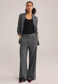 Szürke kockás blézer hozzá illő, bő szárú nadrággal. Fekete, testhez simuló trikó alatta. Figyelemfelkeltő, hegyes bordó cipők és oldalzsebek.