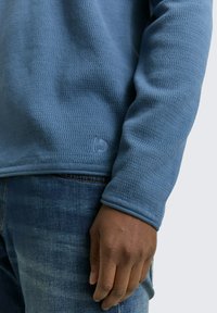 Blauses strukturiertes Langarmshirt mit Rundhalsausschnitt. Verfügt über ein dezentes Markenlogo nahe dem Saum und wird mit dunklen Denim-Jeans kombiniert.