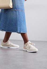 Sneakers sportive beige con tomaia texture e lacci, abbinati a una gonna midi in denim blu chiaro. Superficie liscia su un pavimento grigio.