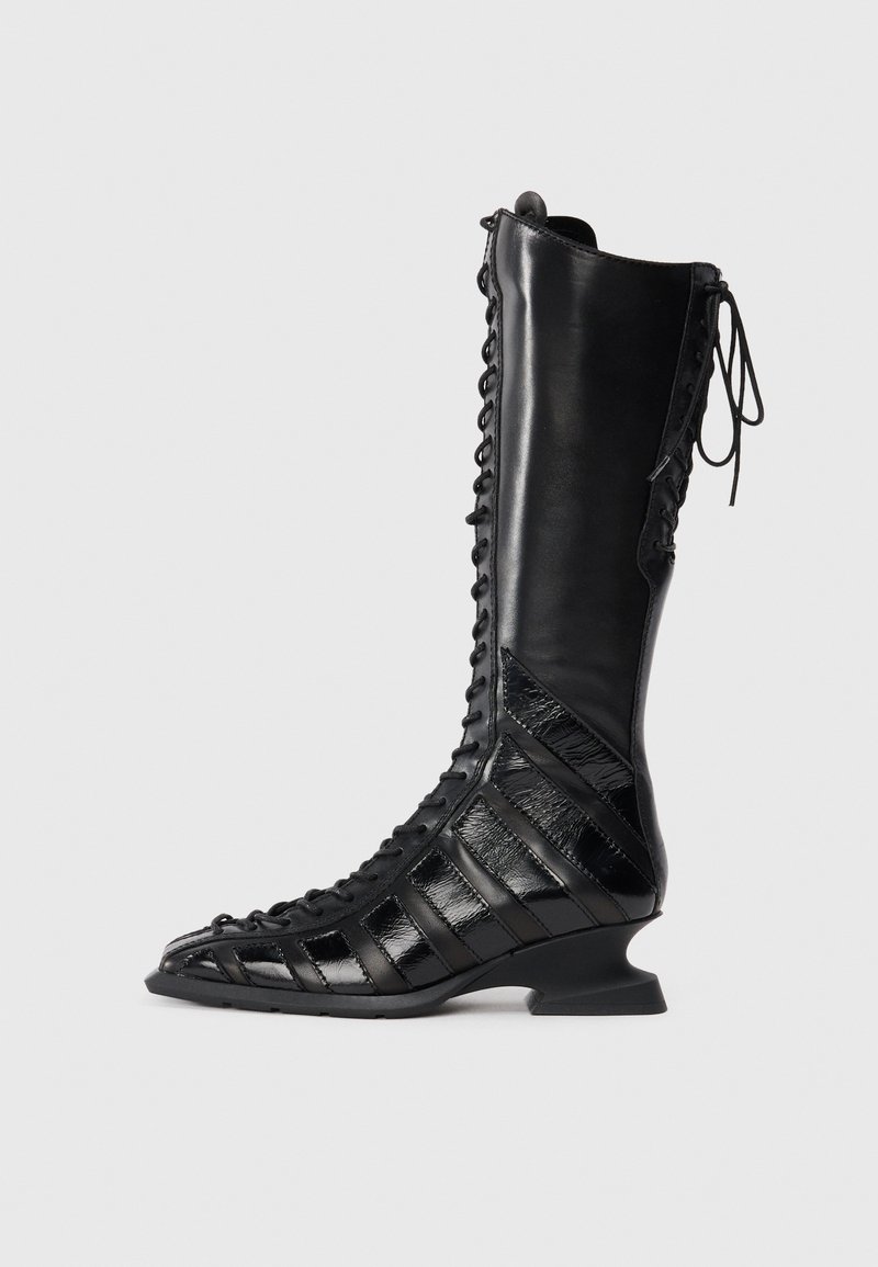 Black knee-high boot with a lace-up front, made of smooth and textured leather, featuring a sculpted heel and geometric panel design.