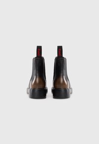 Une paire de bottes Chelsea en cuir noir et marron avec des semelles noires et des languettes rouges "CAMPER", vues de l'arrière sur un fond blanc.