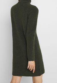 Sidoprofil av en person som bär en mörkgrön, stickad långärmad polotröjdress, som står mot en enfärgad vit bakgrund.