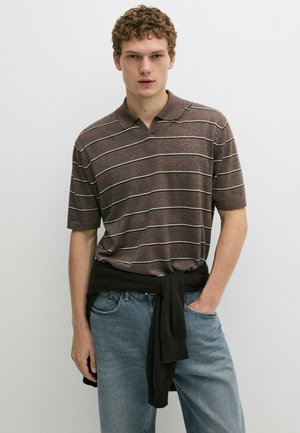 Giovane uomo con capelli ricci che indossa una polo marrone a righe e jeans azzurri, con un maglione nero legato in vita.