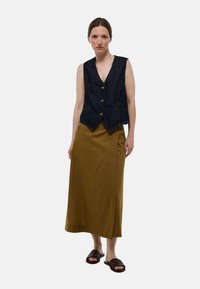Veste en denim avec un décolleté en V et trois boutons, associée à une longue jupe olive avec une fente sur le côté et des sandales brunes à enfiler.