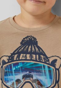 Beige T-shirt met een afbeelding van een beer met een blauwe en paarse bergscène weerspiegeld in zijn skibril. Zacht katoenen materiaal.