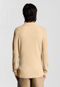 Beige långärmad tröja med en texturerad yta, med en avslappnad passform och smal krage. Sömlösa axlar och ribbade muddar.