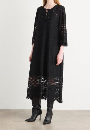 Femme vêtue d'une robe midi en dentelle noire à manches trois-quarts et de bottes noires pointues à talons hauts, debout sur un sol uni.
