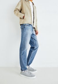 Jasnobeżowa kurtka z kołnierzem, biała koszulka, niebieskie dżinsy oraz szare zamszowe trampki z białymi podeszwami, prezentowane na jasnym tle.