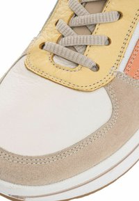 Zapatilla blanca con acentos de ante beige y un revestimiento de cuero amarillo. Presenta cordones planos grises y costuras visibles a lo largo de los bordes.