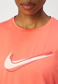 Camiseta deportiva de manga corta en color coral con cuello redondo y logo de Nike en blanco y rosa en el pecho. Tejido suave y que absorbe la humedad.