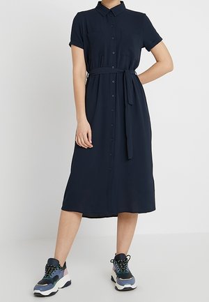 Kobieta ubraną w granatową, zapinaną na guziki sukienkę midi z krótkimi rękawami i paskiem w talii, w połączeniu z wielokolorowymi, masywnymi sneakersami.
