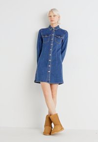 Μπλε τζιν φόρεμα με κουμπιά μπροστά, γιακά και τσέπες στο στήθος; συνδυασμένο με καφέ μποτάκια μέχρι τον αστράγαλο, με λεία υφή και μήκος μέχρι το γόνατο.