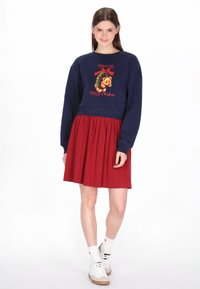 Tengerészkék pulóver "Boldog Karácsonyt" felirattal és medve grafikával, piros pliszírozott szoknyával párosítva. A modell fehér zoknit visel fekete részletekkel és sportcipőt.