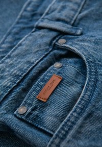 Camisa de mezclilla en azul oscuro con textura visible, botones plateados y una etiqueta de cuero marrón cosida. Presenta un cuello y bolsillos.