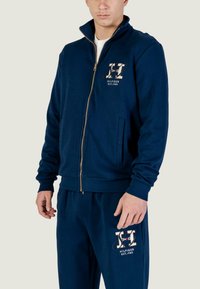 Giacca blu navy con zip e logo dorato, dotata di tasche laterali e polsini a costine. Pantaloni jogger coordinati blu navy con dettaglio di marchio abbinato.